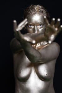 Shiny gold-painted woman with outstretched hands, black background, artistic portrait.
