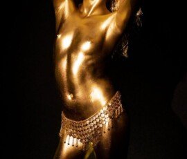 Glamorous woman covered in gold body paint poses with confidence against a dark backdrop. Shiny gold body paint beauty shot with gold coin belt accessory on a black background.