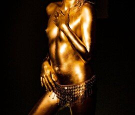 Close-up of a woman covered in gold body paint wearing a belly dance belt against black background. Shiny gold body paint woman with belly dance belt on black background.