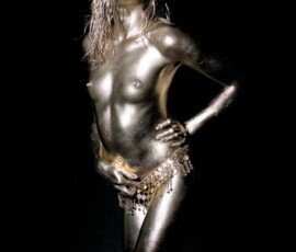 Glamorous model covered in metallic gold body paint with jewelry, creative photo shoot. Shiny metallic body paint on model with gold jewelry, artistic photo shoot with dark background.