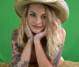 Portrait of a confident woman with tattoos, smiling, and wearing a straw hat against a green background. Cute woman with tattoos wearing a wide-brimmed straw hat in front of a green screen.
