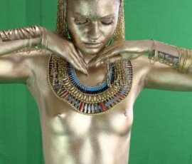 A woman covered in gold body paint, wearing Egyptian-style jewelry and headpiece, with a green screen backdrop for photo shoot. Golden body paint and Egyptian-inspired jewelry on a woman with a green background.