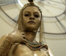 A striking golden statue of a woman adorned with tribal jewelry, standing confidently against a background featuring a large clock. Golden statue of a woman with tribal jewelry and headpiece, standing indoors with a large clock background.