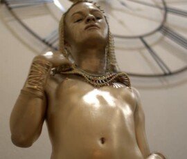 Elegant golden statue representing cultural art with jewelry, showcased in a stylish indoor setting. Gold-painted classical sculpture of a woman with jewelry, against a clock background, artistic and contemporary photography.