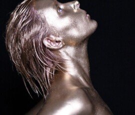 Glamorous woman with metallic gold body paint, wet hairstyle, and dark background in artful fashion photography. Glossy metallic gold body paint on woman with wet hair, dark background, artistic fashion photography.