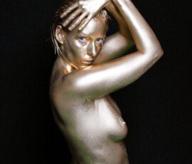 A woman covered in metallic gold body paint, posing with hands on her face and body, artistic makeup, studio shot for creative photography. Shiny gold body paint on a woman with striking pose and dramatic lighting against black background.