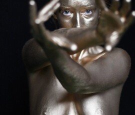 Gold-painted female model with intense gaze, reaching out hands, black backdrop. Shiny gold-painted woman with outstretched hands, black background, artistic portrait.