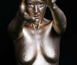 A woman covered in reflective gold body paint, reaching out with her hands and gazing intensely at the camera, against a dark background. Shiny gold body paint woman with intense gaze reaching out towards the camera.