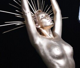 Elegant metallic woman sculpture with raised arms and radiant design, representing strength and beauty in modern art exhibitions. Shiny metallic sculpture of a woman with arms raised, golden rays emanating from her head, art installation, contemporary sculpture, modern art piece.