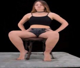 Young woman sitting confidently on a black chair with a black background, wearing a crop top and shorts, showcasing modern fashion style. Seated woman in black crop top and shorts on black chair against black background.